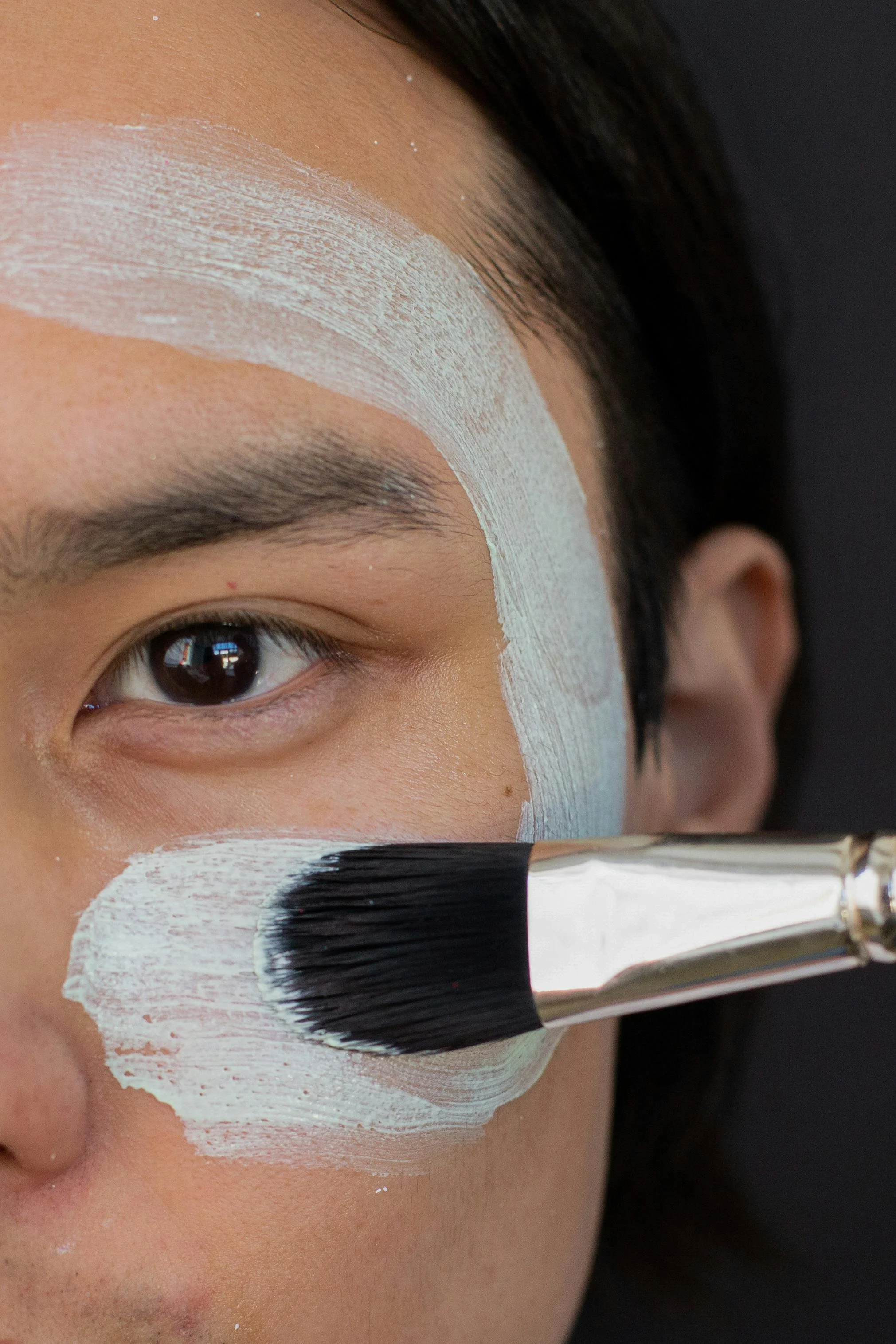Close-up of a person with makeup paint on their face, applying white paint with a large makeup brush near their cheek and eyebrow.