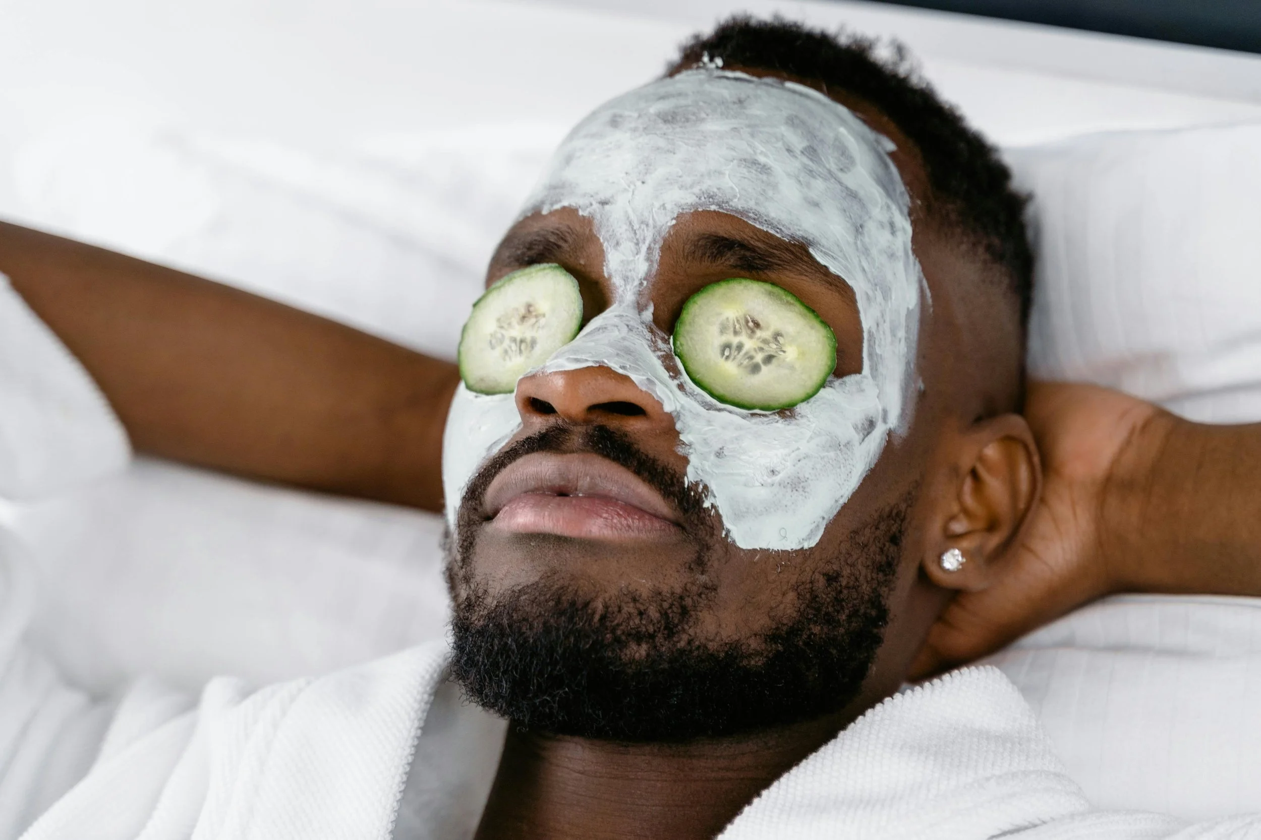 A man with a white facial mask and cucumber slices over his eyes, relaxing with his arms behind his head, lying on a white pillow.
