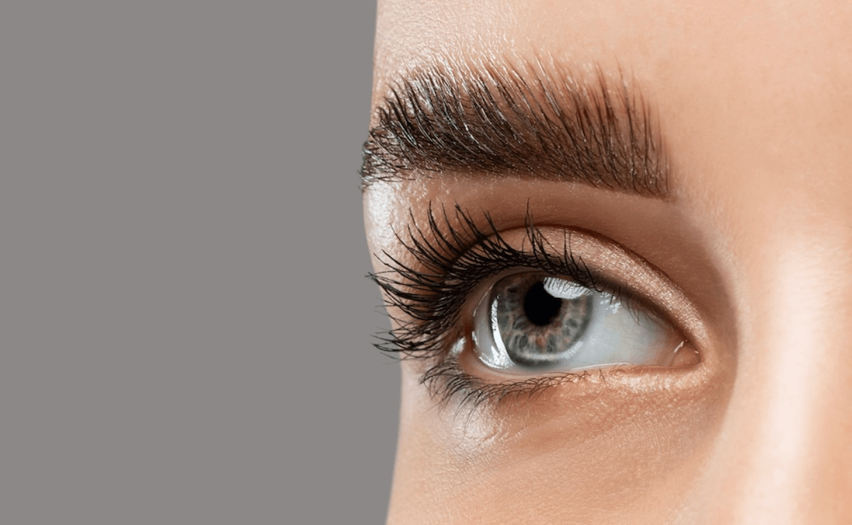 Close-up of a person's eye with detailed eyelashes, eyebrow, and skin texture against a gray background.