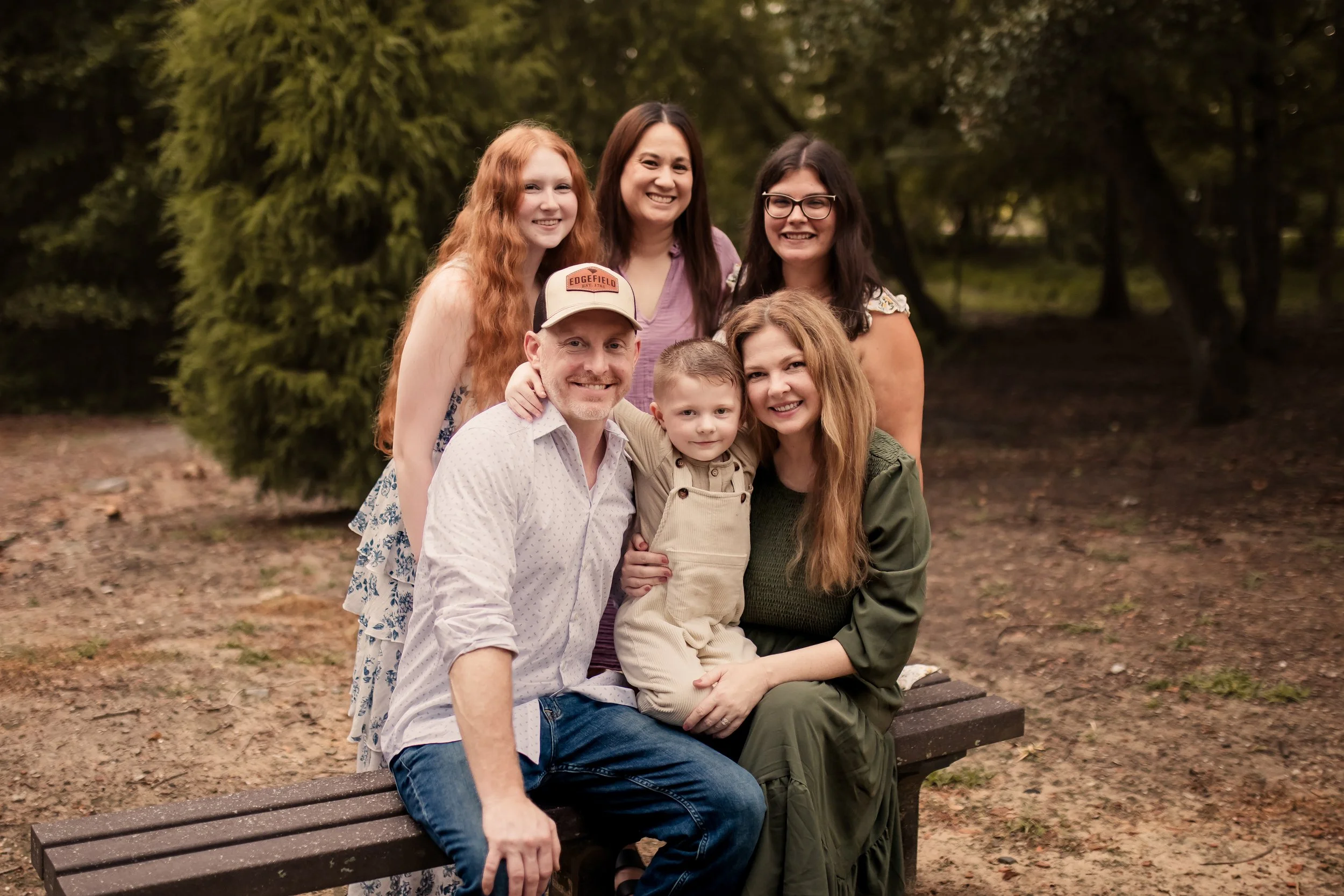 A group of seven people, including children and adults, sitting and standing on a bench outdoors surrounded by trees. They are smiling and posing for a photo.