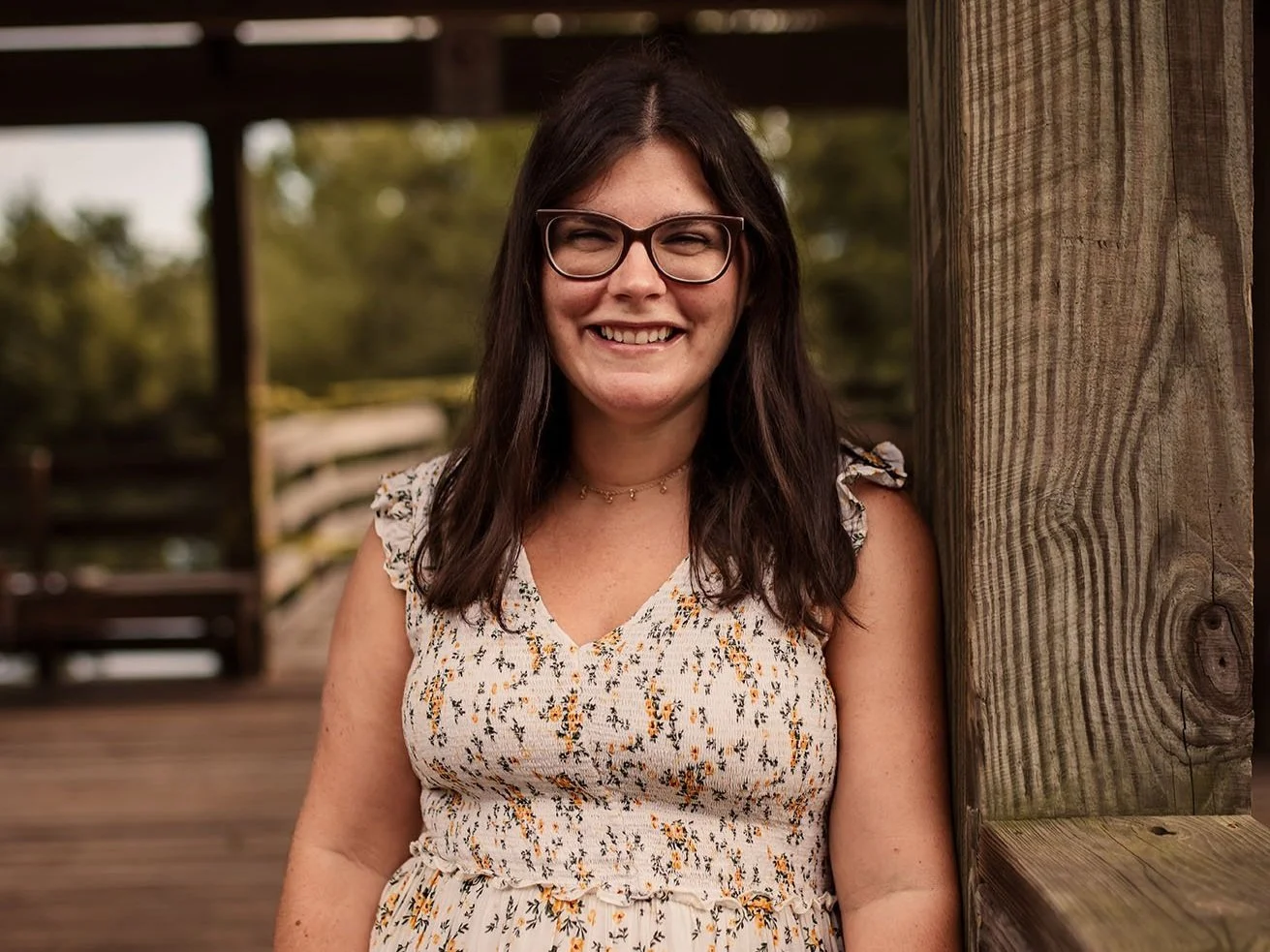 A smiling woman with glasses and dark brown hair standing outdoors next to a wooden post on a bridge.