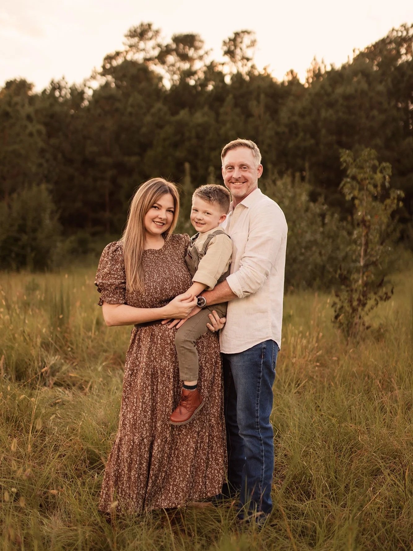 A family of three standing outdoors in a grassy field during sunset, smiling at the camera.