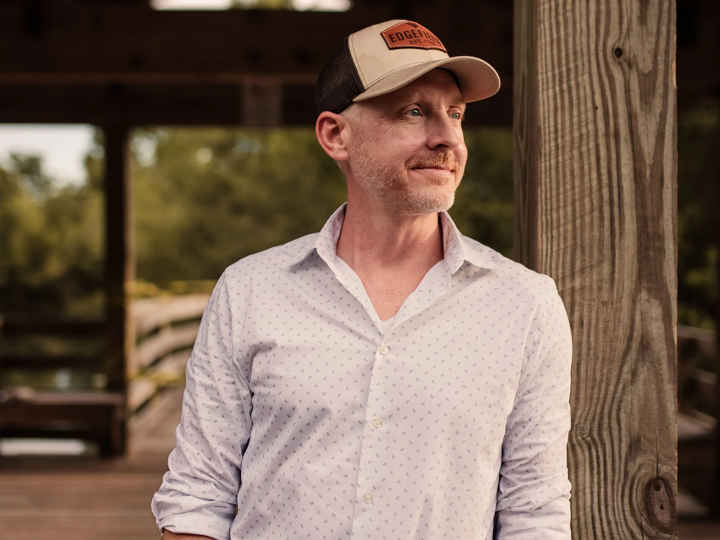 A man with a beard and mustache wearing a white shirt with small blue patterns and a cap with an orange patch, standing outdoors under a wooden structure, looking to the side with a slight smile.