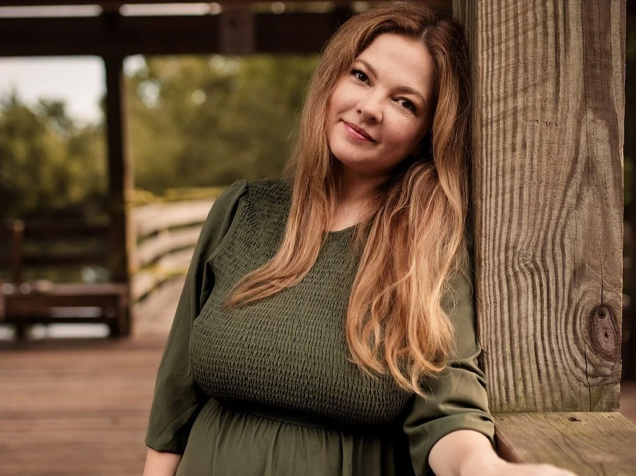 A woman with long, wavy, reddish-blonde hair leaning against a wooden post outdoors. She is wearing an olive-green top and smiling at the camera. The background features a blurred outdoor setting with greenery and wooden structures.