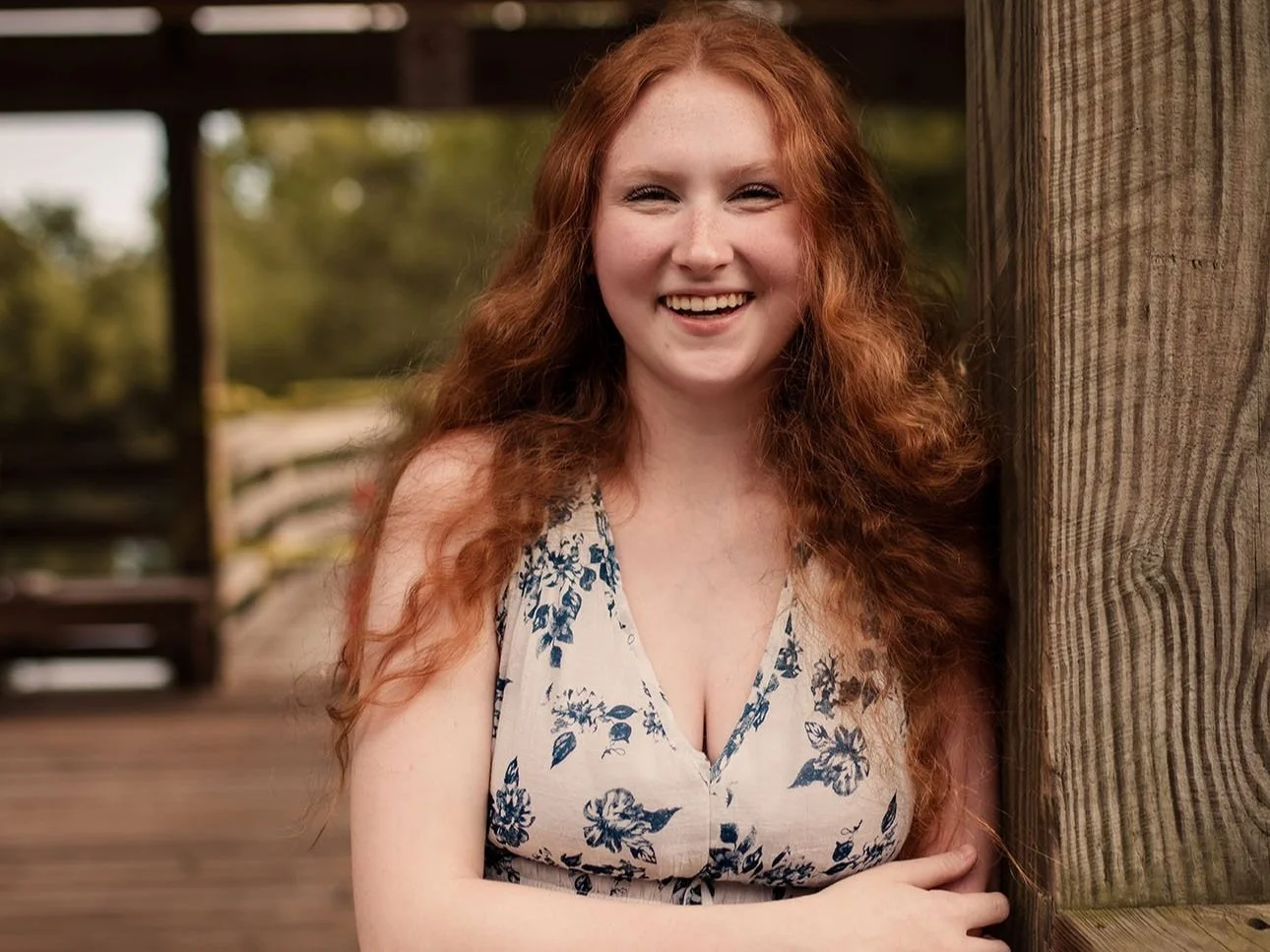 A smiling woman with long, curly red hair standing outdoors near a wooden post, wearing a sleeveless floral dress.