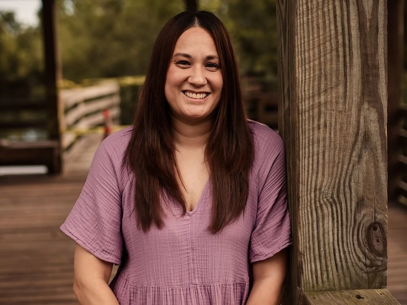 A woman with long brown hair, smiling, standing next to a wooden post outdoors during daytime.