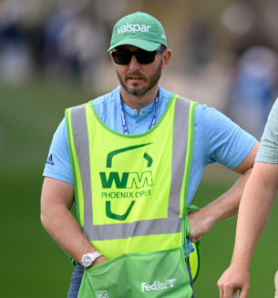 A man wearing a green cap and sunglasses, with a yellow vest over a blue shirt, standing outdoors at a golf event.