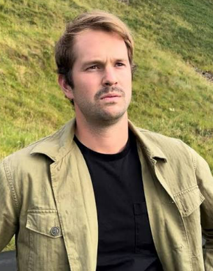 A man with light brown hair and a serious expression, wearing a beige jacket and black shirt, standing outdoors on a grassy hill.