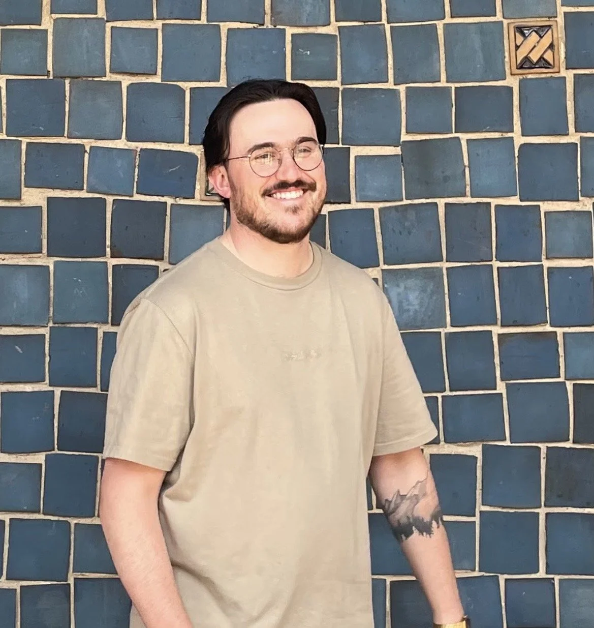 A man with glasses and a beard smiling, standing against a dark brick wall with a small decorative vent in the upper right corner.