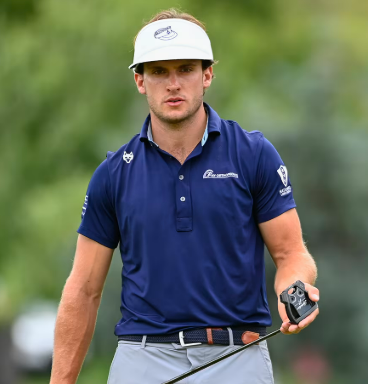 A male golfer wearing a white cap and navy blue shirt, holding a golf club, on a golf course with greenery in the background.
