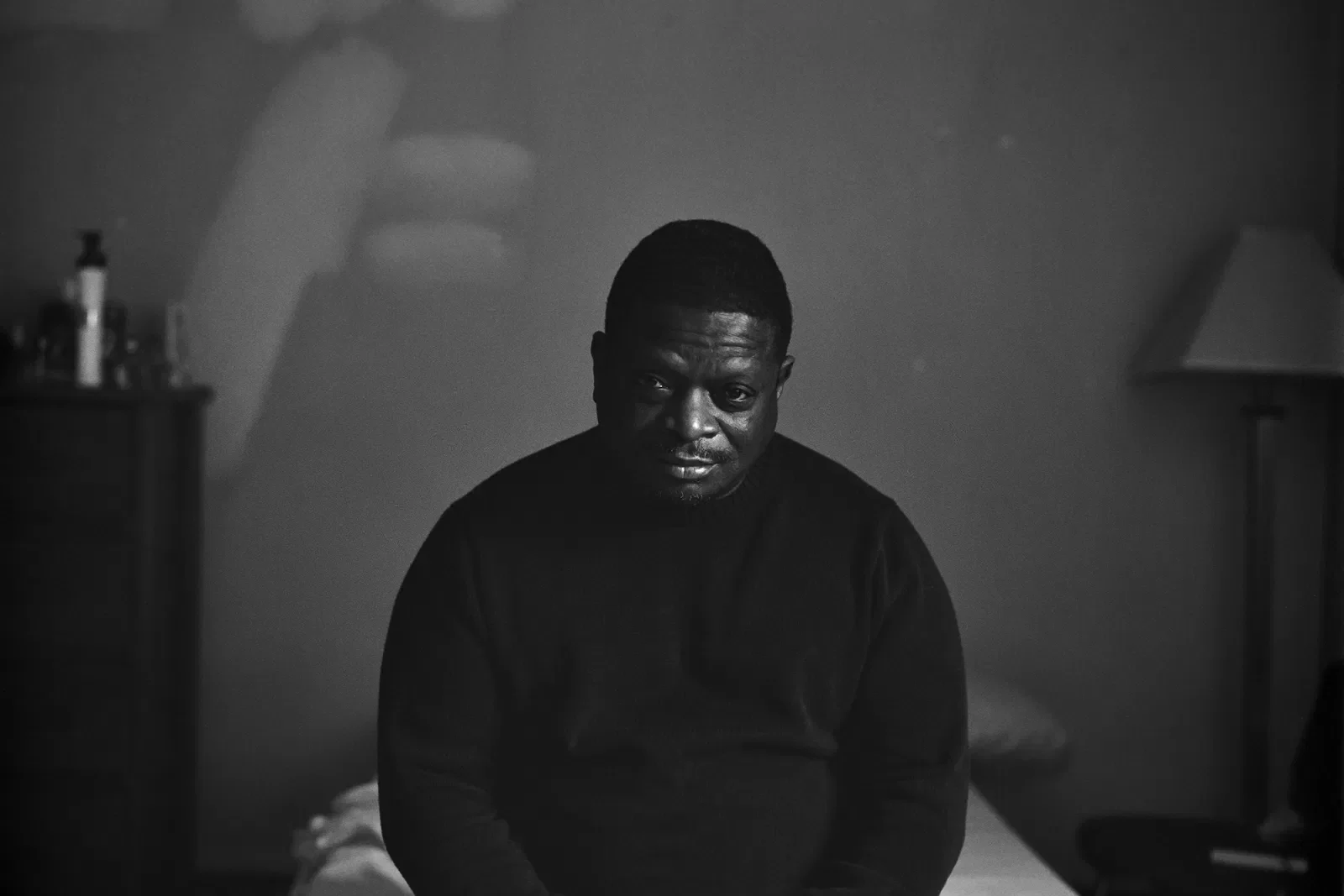Black and white portrait of a man sitting in a dimly lit bedroom, captured by a storytelling agency for nonprofits to highlight social impact and purpose-driven narratives
