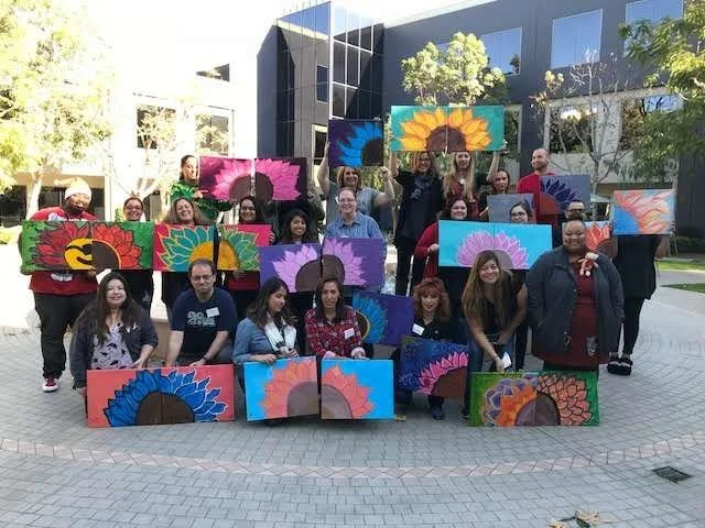 Group of people holding paintings of colorful sunflower designs outdoors.