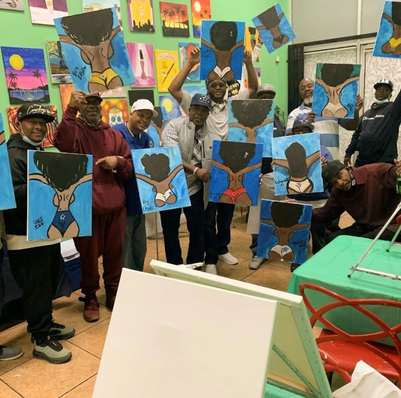 Group of people holding paintings of a woman with natural hair and arms outstretched, inside an art studio with colorful paintings on the wall.
