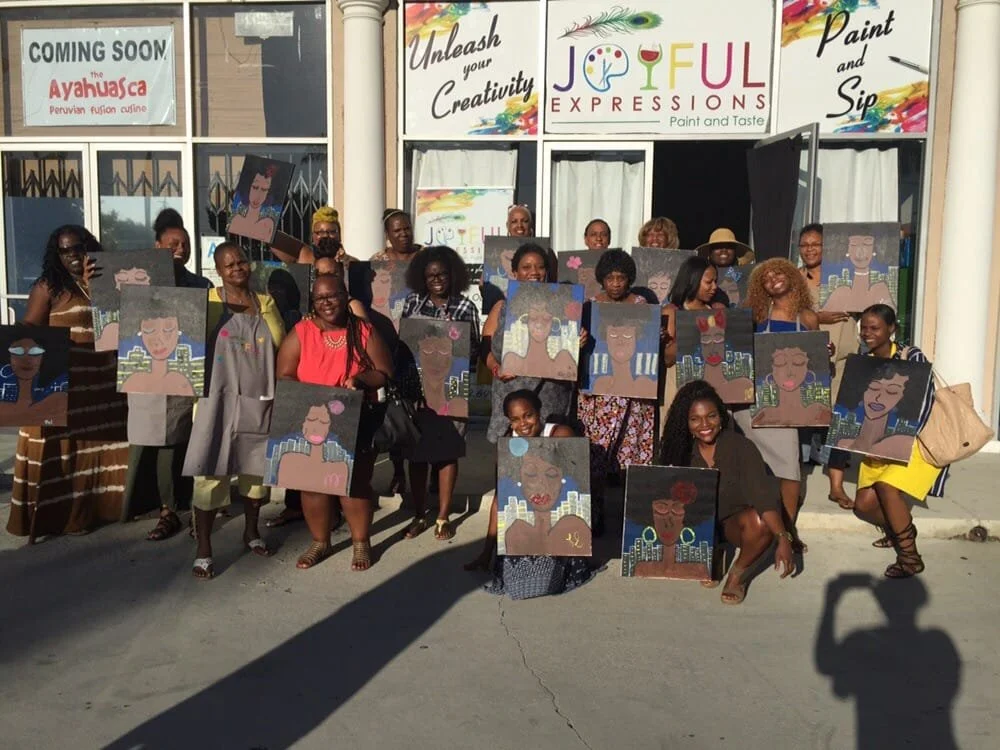 Group of women holding paintings of women with various hairstyles and accessories outside a paint and sip studio.