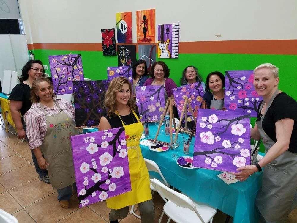 Group of women participating in a painting class, holding up their completed purple floral paintings featuring cherry blossoms, with additional artwork displayed on the colorful wall behind them.