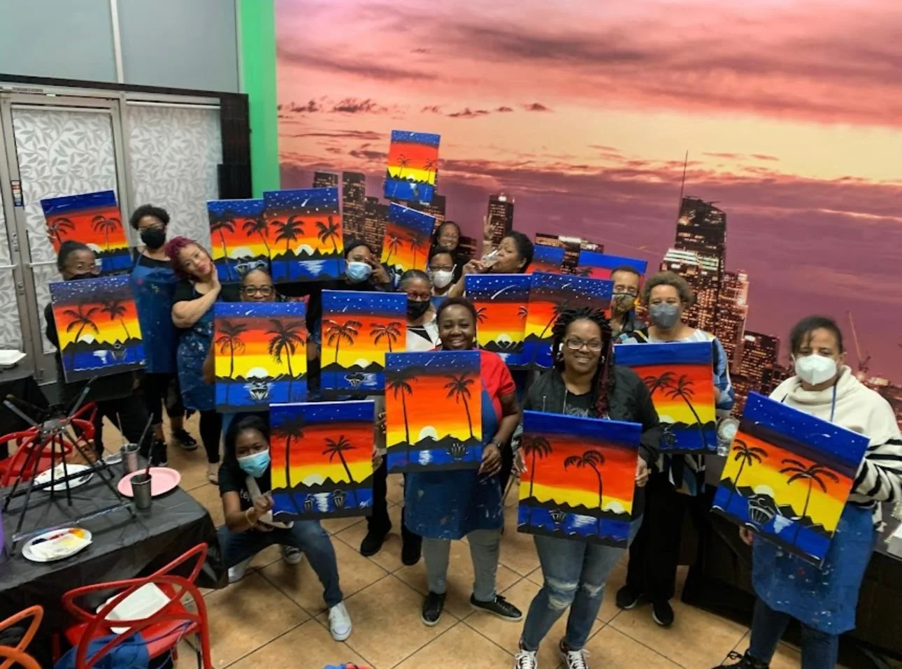 A group of people holding canvas paintings of a sunset with palm trees and a city skyline background, in an indoor art studio with a large sunset cityscape mural.