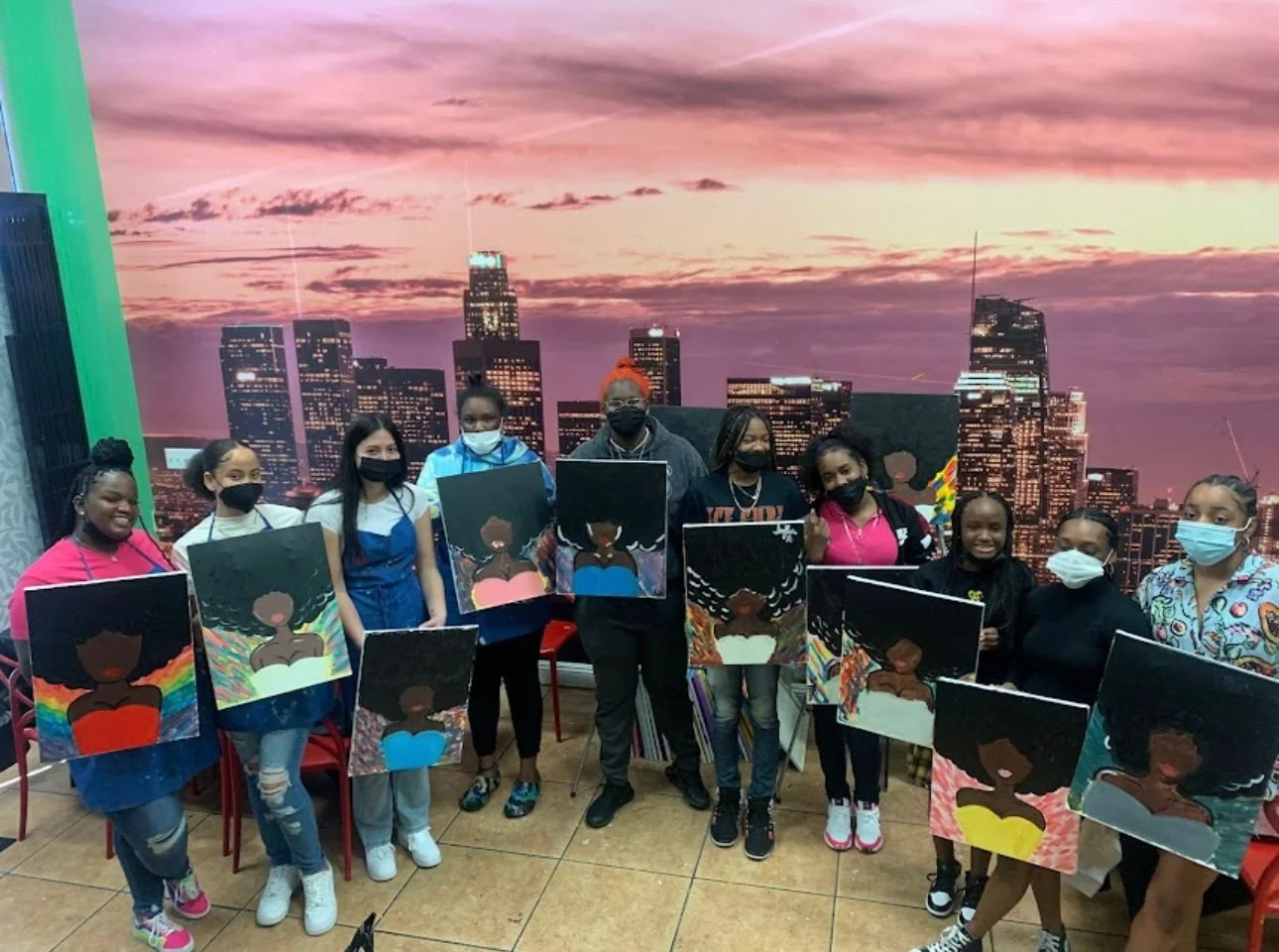 A group of young artists, mostly girls, holding paintings of a woman with afro hair, standing in front of a cityscape backdrop at an indoor art event.