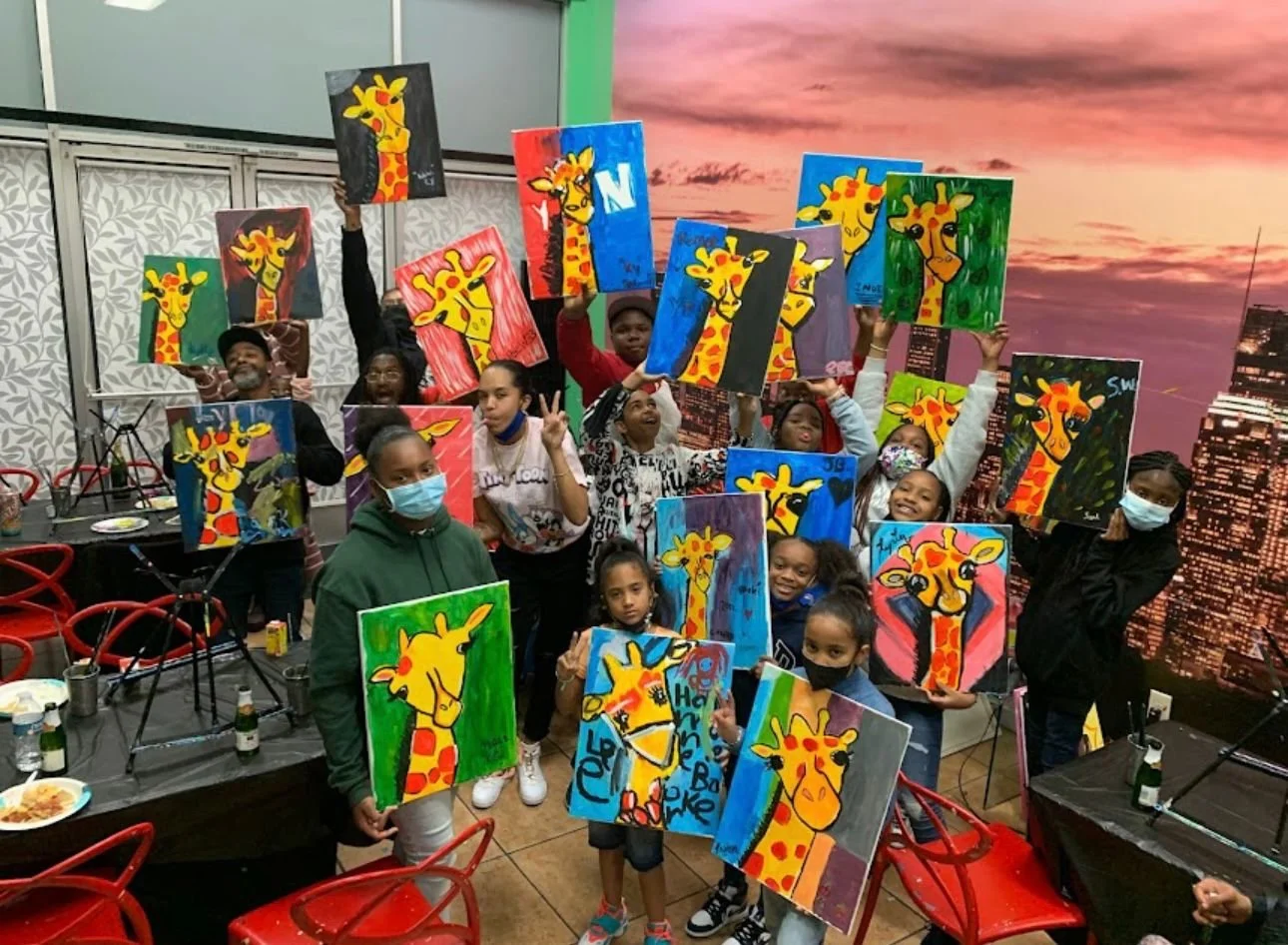 Group of children and adults at an art class holding up colorful paintings of giraffes, some wearing masks, with a city skyline mural in the background.