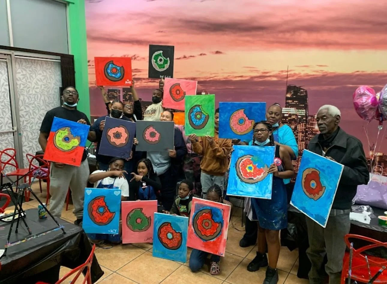 Group of people holding colorful abstract paintings at an indoor art event with a city skyline backdrop.