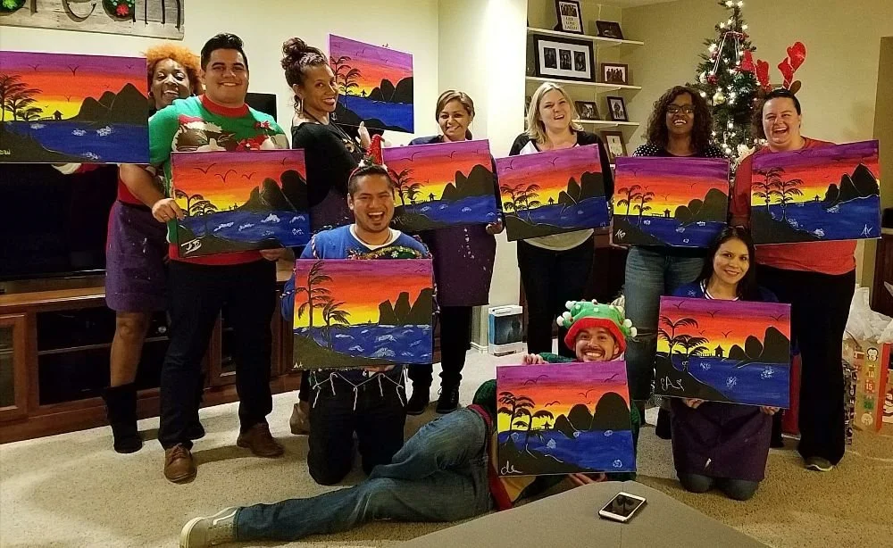 Group of people at a holiday party holding paintings of sunset landscapes with trees and mountains. Some are wearing Christmas-themed attire, with one person wearing a holiday hat and others sporting reindeer antlers.