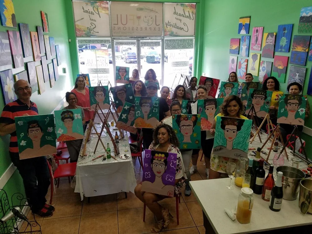 A group of people at a painting class displaying their portraits of a woman with flowers in her hair inside a colorful art gallery.