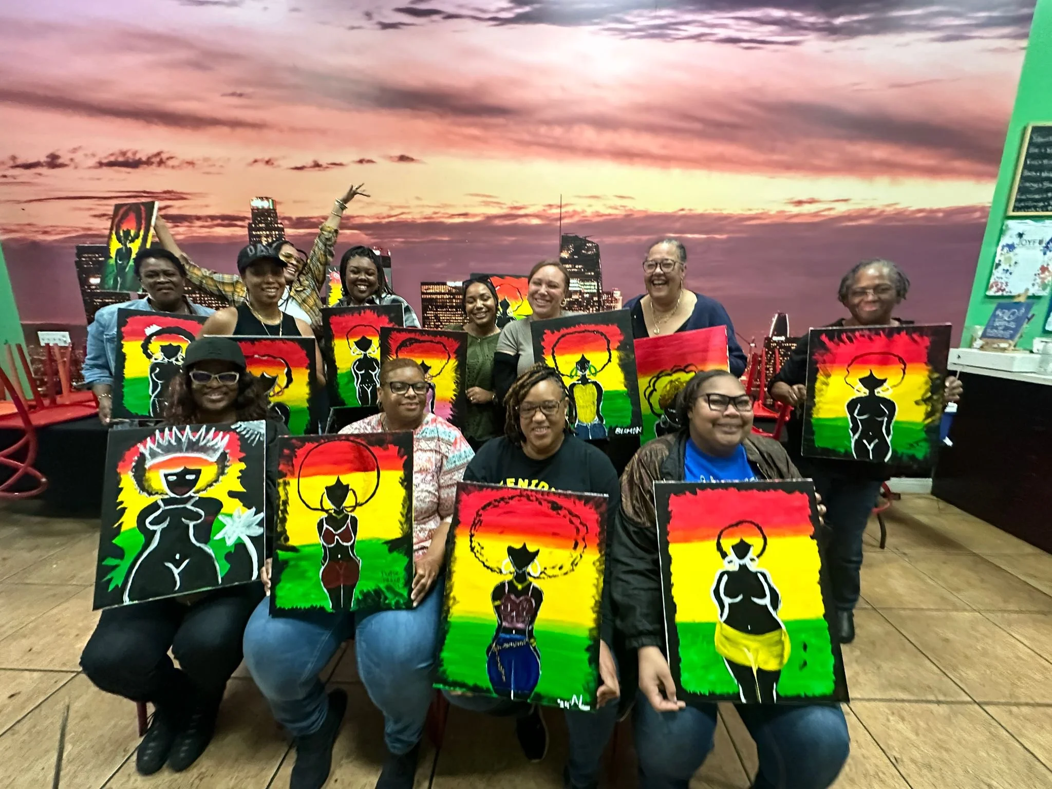 Group of women holding colorful paintings depicting stylized black female figures with sun or halo backgrounds, inside a room with a cityscape sunset mural.