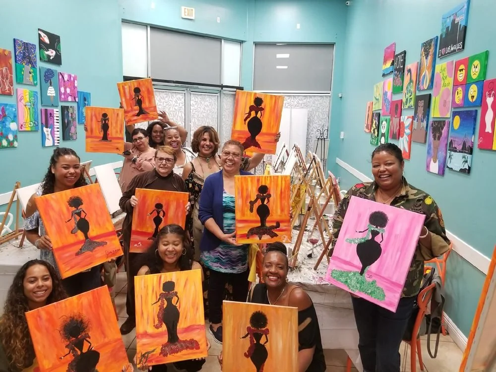 A group of women in an art studio holding paintings of a woman with an afro hairstyle in silhouette against a colorful sunset background. The women are smiling and the studio is decorated with various colorful artwork on the walls.