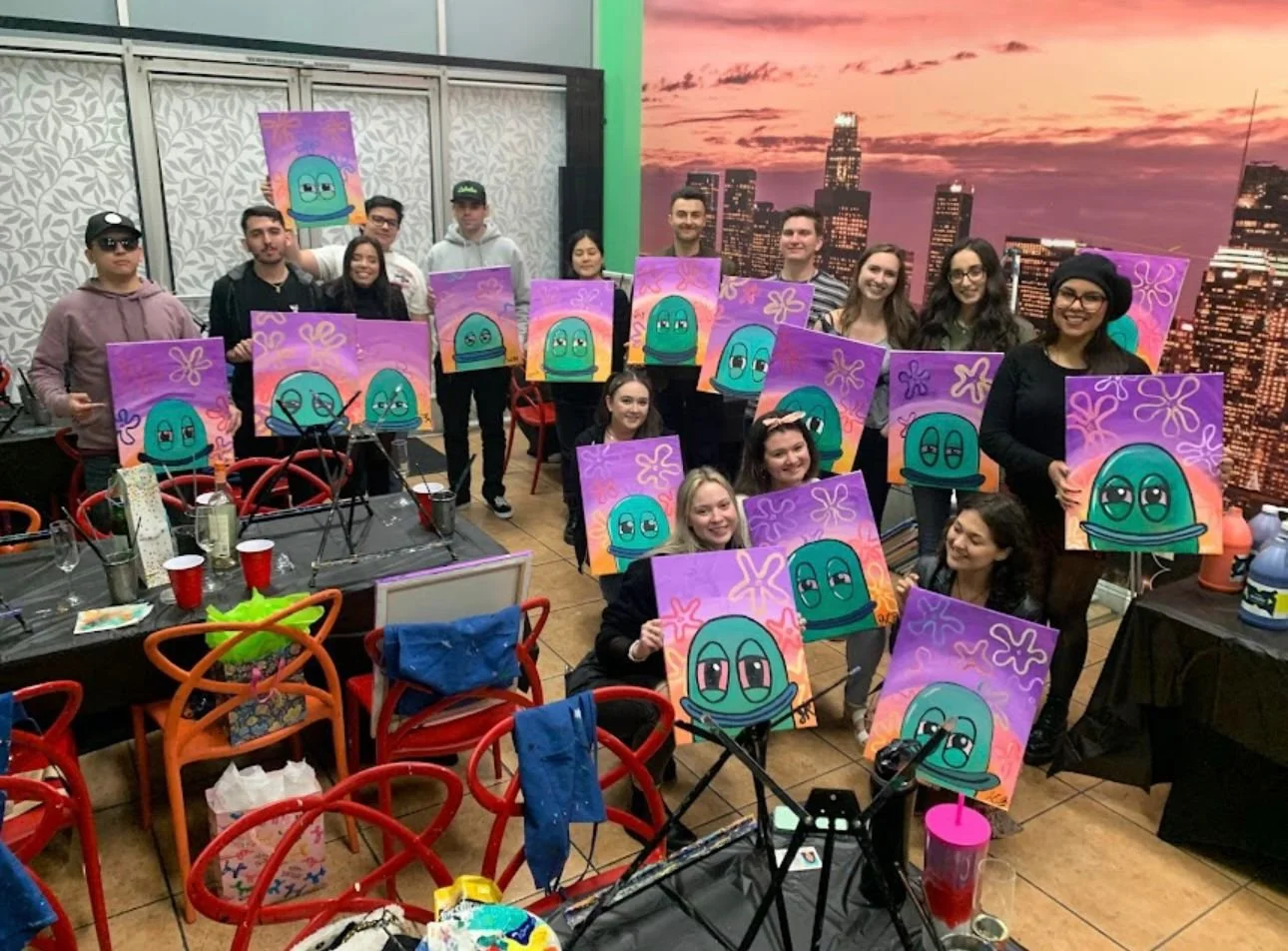 A group of people showing their paintings of cartoon jellyfish with pink, purple, and orange backgrounds at an indoor art event.