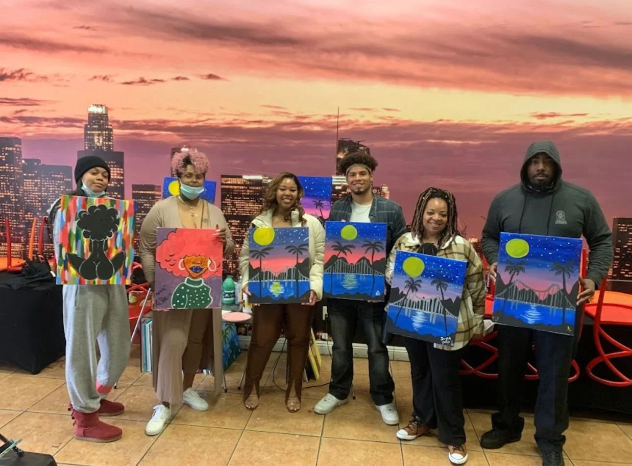 Group of six people holding completed landscape paintings in front of a city skyline mural at sunset.