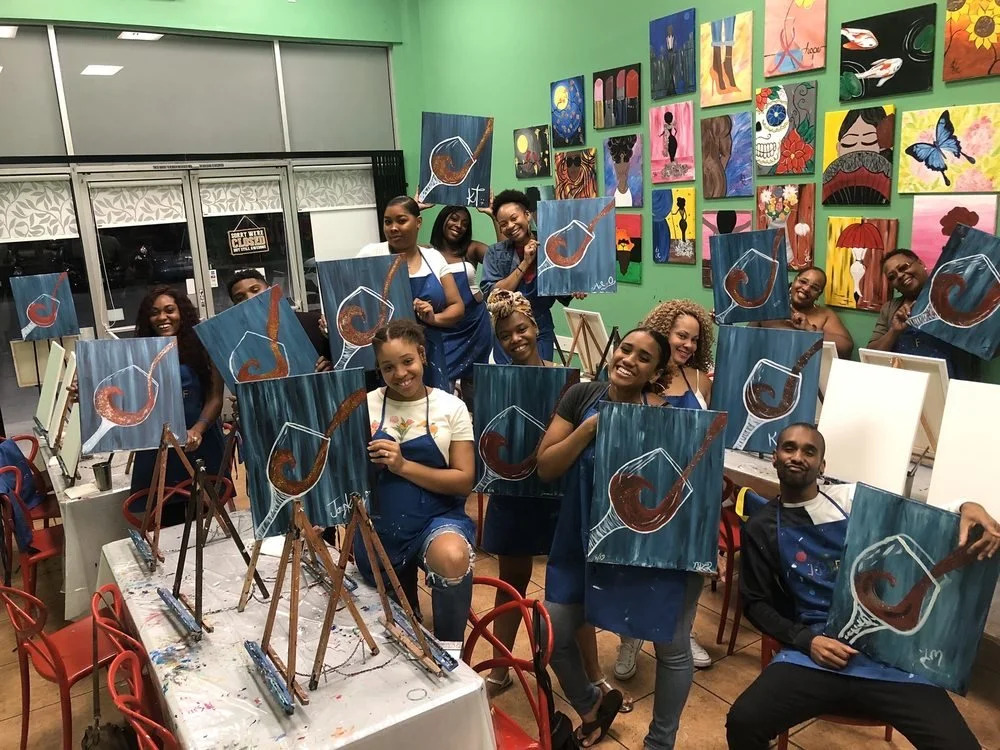 A group of people at a painting class holding up their completed abstract paintings of a bowl with a spoon, with many colorful artworks displayed on the wall behind them.