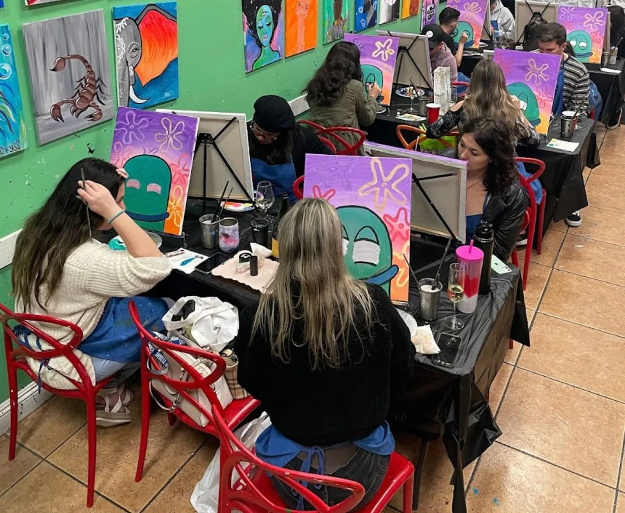 People participating in a painting class creating colorful artwork featuring cartoon-style faces and abstract designs in a casual indoor setting.