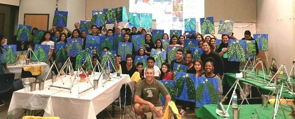 Group of children and adults in a classroom holding up artwork of abstract blue and green paintings, with tables and art supplies in the foreground.