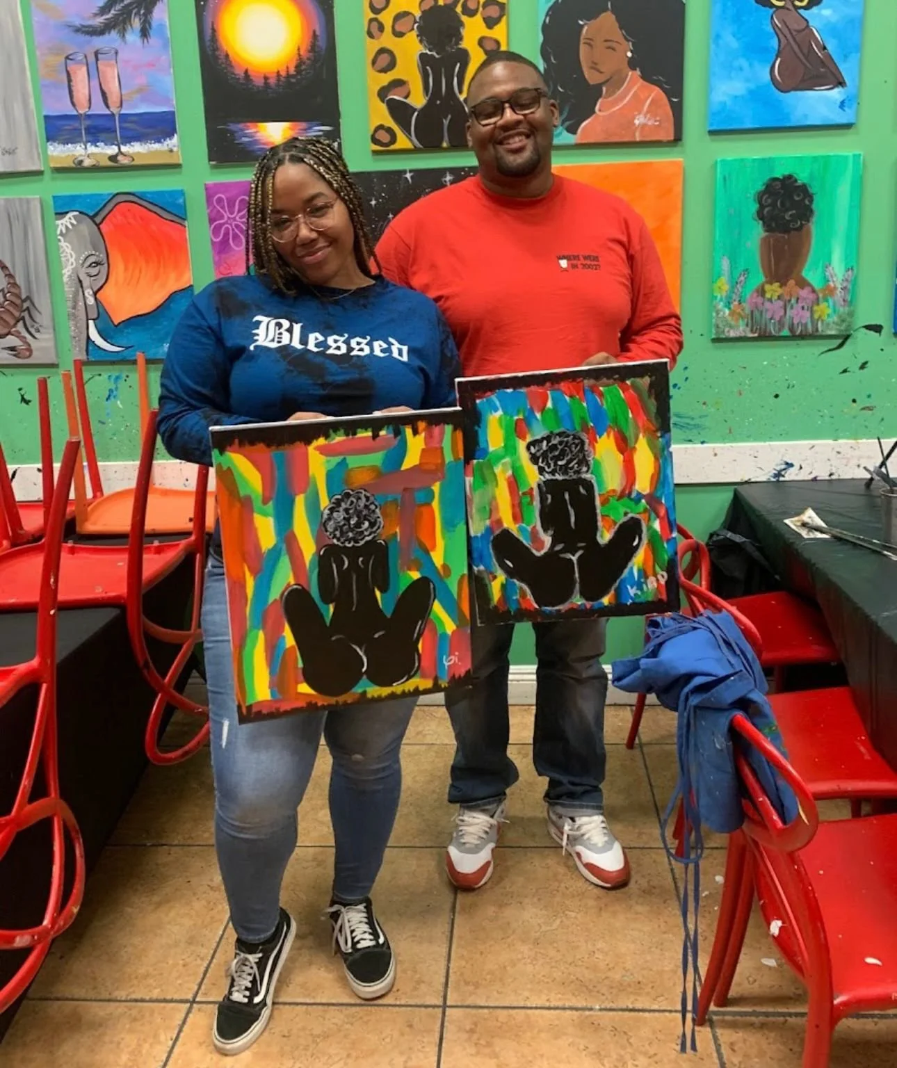 Two people standing in an art studio holding paintings of a black silhouette of a seated child with drying hair, set against colorful abstract backgrounds. The studio walls are decorated with various paintings, including a sunset, a woman, and animal