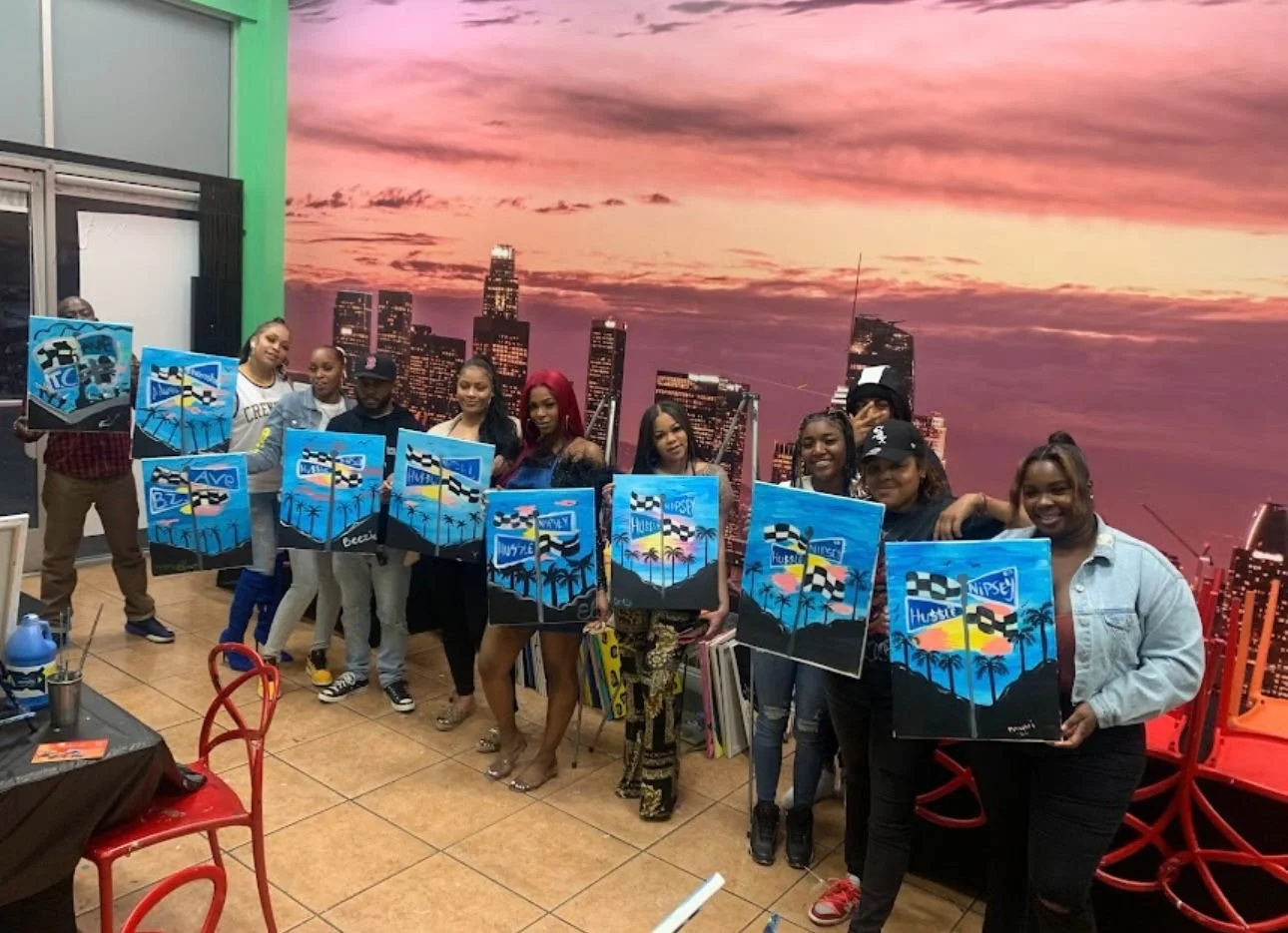 A group of ten women standing indoors in front of a mural of a city skyline at sunset, each holding a painting of a beach scene with palm trees and a sunset, smiling for the photo.