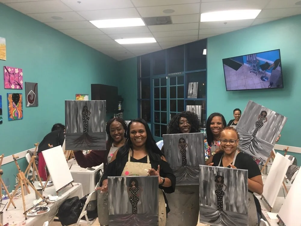 Group of women at a painting class holding their finished paintings of a woman in a polka-dot dress.