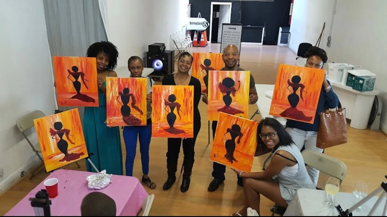 Group of six African American women and one African American man smiling and holding up completed paintings of a silhouette of a woman against a bright, fiery orange and yellow background, in an indoor art class or workshop setting.