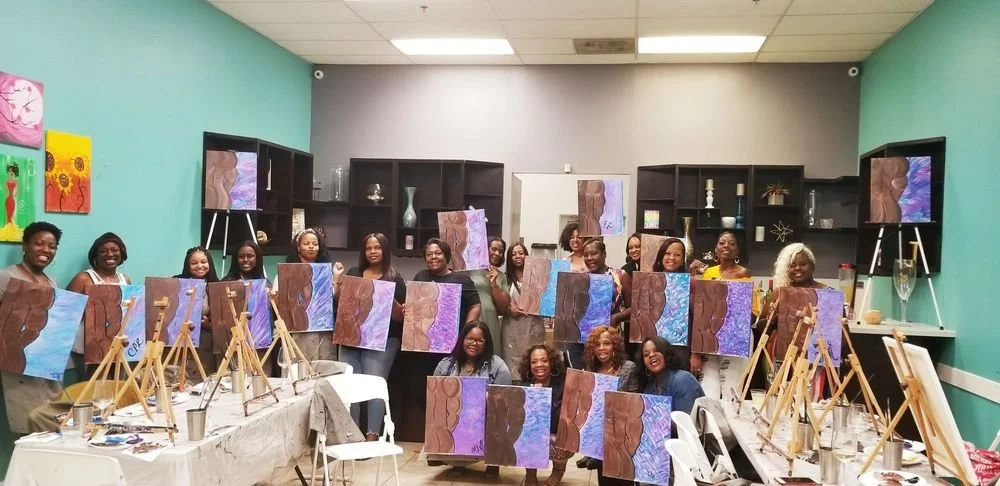 Group of women holding up completed abstract paintings in a classroom or art studio.