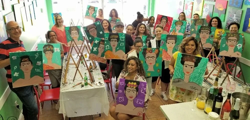 Group of people in a classroom holding up their individual painted portraits of Frida Kahlo, with some standing behind easels and others sitting or standing, surrounded by colorful artwork on the walls.