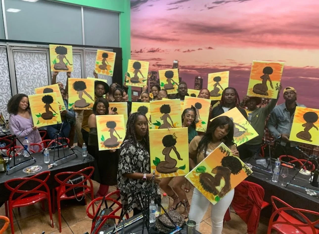 Group of people at an art class holding paintings of a silhouette of a woman with natural hair, against sunset backgrounds. Some participants are smiling, and the room is decorated with a sunset mural and red chairs.