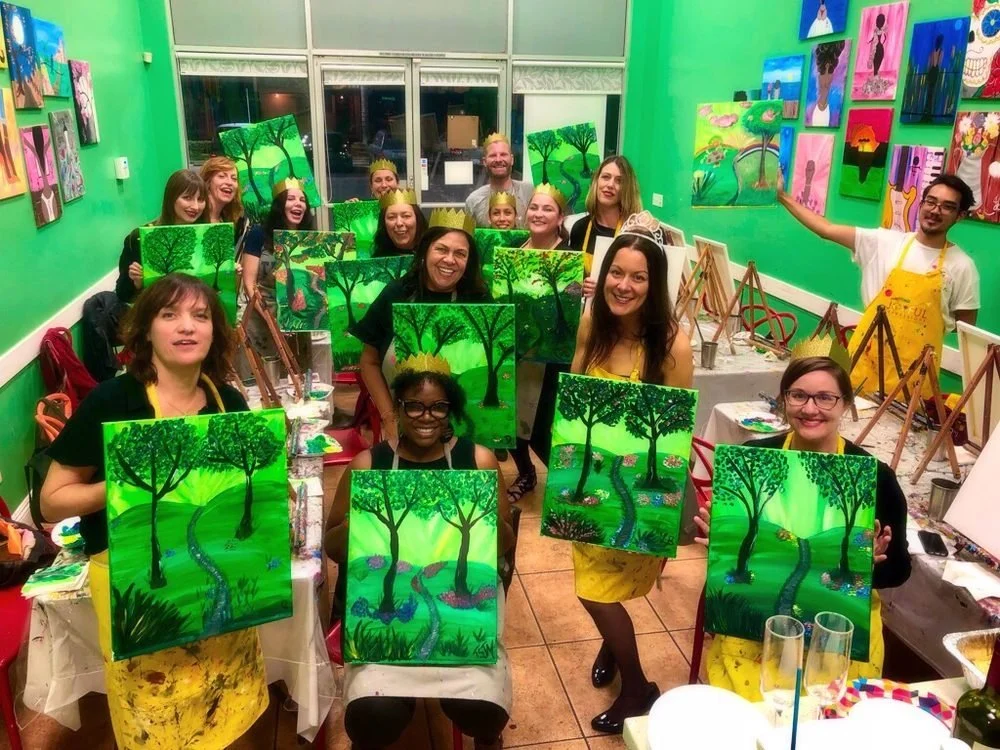 Group of people participating in a painting class, holding their landscape paintings of a green forest path with trees, inside a vibrant green-walled art studio.