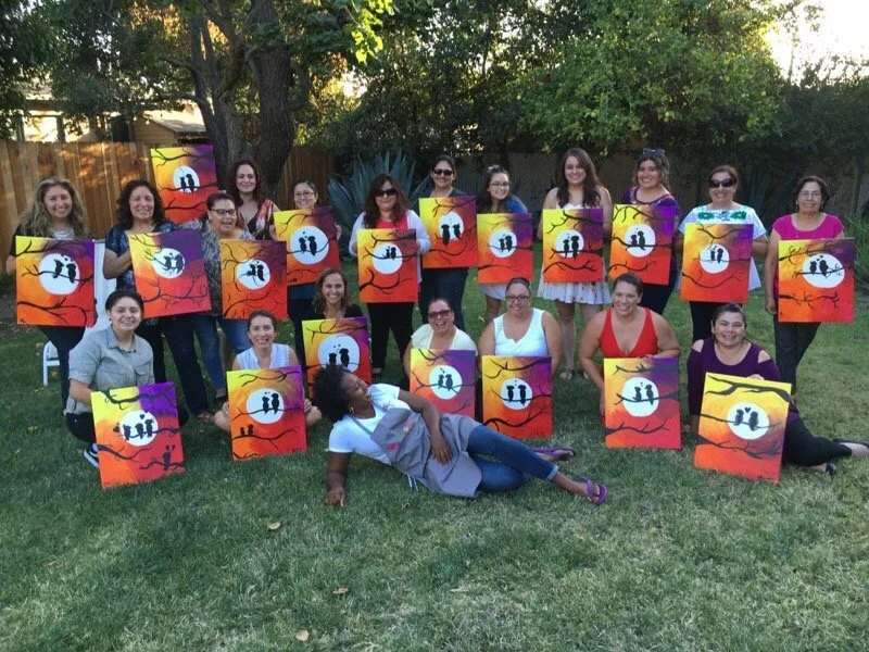 A group of twenty women outdoors in a backyard, holding colorful paintings of a sunset scene with trees and two people holding hands, all smiling.