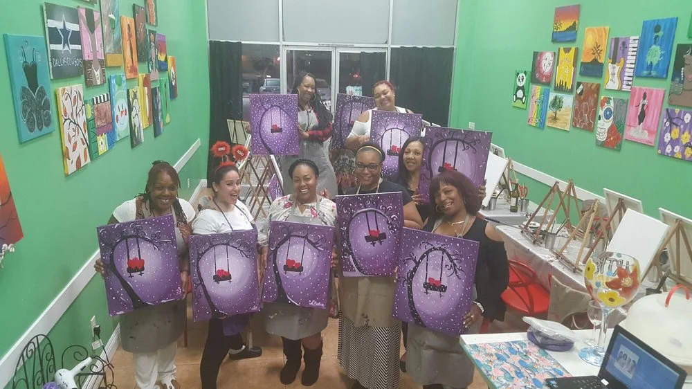 Group of women celebrating a painting class in a colorful art studio. They are holding up purple canvases with a swing painting. The walls are decorated with various colorful art pieces.