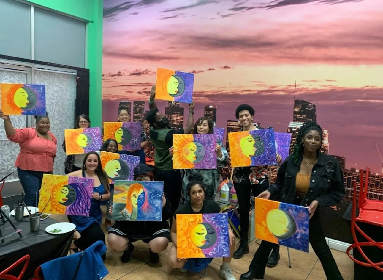 Group of people holding colorful paintings of a sun and moon on canvas, with a cityscape sunset mural on the wall behind them, in an indoor art studio.