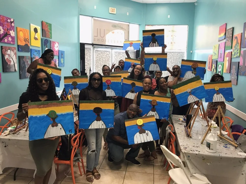Group of smiling women and one man holding up their paintings of a woman with a colorful sky background inside an art studio.