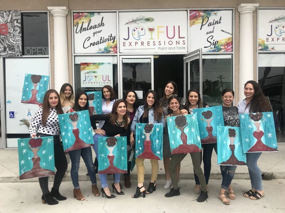 A group of women standing outside an art studio holding paintings of a woman with an updo hairstyle, blue background, and white stars. The studio sign reads "JOFUL EXPRESSIONS Paint and Taste" and has the phrases "Unleash your Creativity" and "Paint