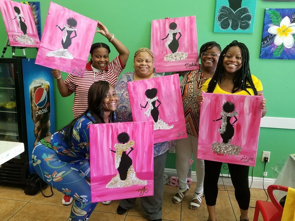 Five women posing together indoors, holding pink canvas paintings of a silhouette of a woman in a dress with a fluffy skirt, against a pink background. The women are smiling and appear to have completed a painting class or workshop.