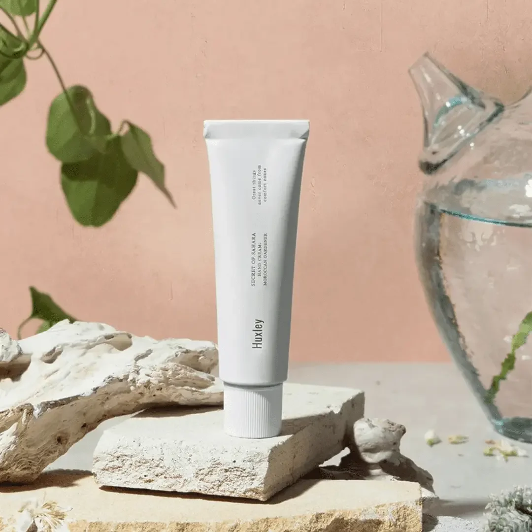 A white tube of Huxley Secret of Sahara moisture cream stands upright on a textured beige stone, surrounded by natural decor including a piece of driftwood, a glass vase with water and greenery, and small white flowers, against a pink wall background.
