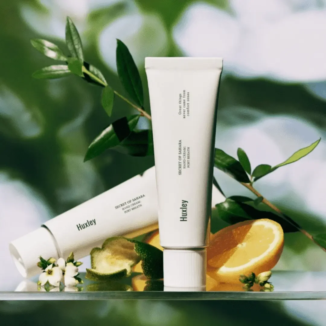 Two white tubes of skincare product labeled Huxley Secret of Sahara, surrounded by lemon slices, a small white flower, and green leaves on a reflective surface with a blurred green leafy background.