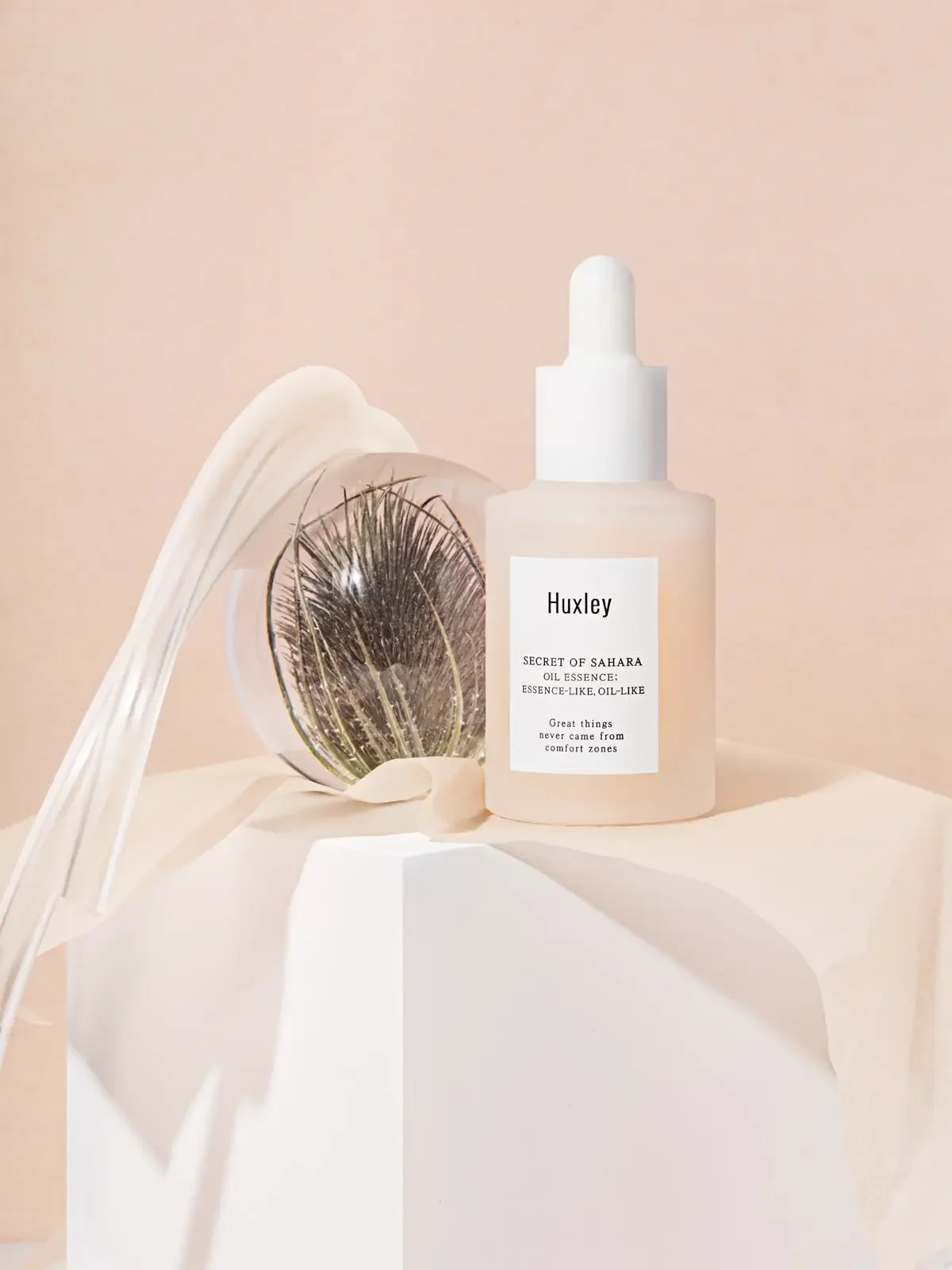 A skincare product bottle labeled 'Huxley' with a dropper cap, alongside an open jar of eye cream with a single false eyelash, all set against a soft pink background.