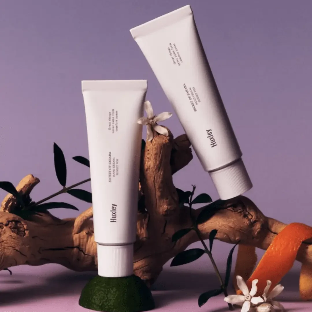 Two white tubes of skincare products labeled "Huxley" placed among a lemon, a lime, an orange peel, spices, and a branch with leaves and white flowers against a purple background.